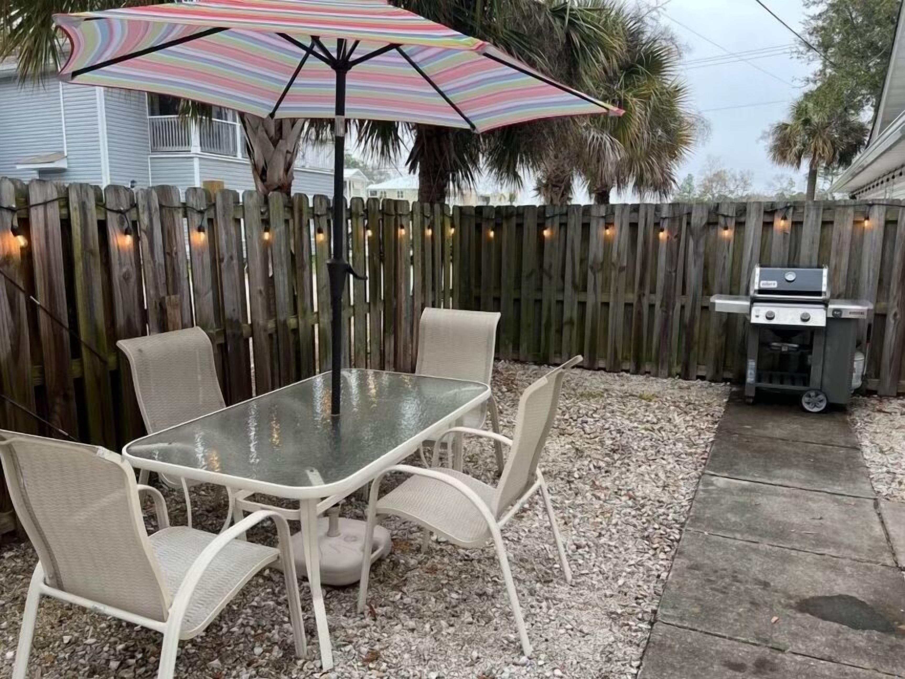 A patio with a glass table and four chairs, a striped umbrella, a wooden fence, and a barbecue grill.