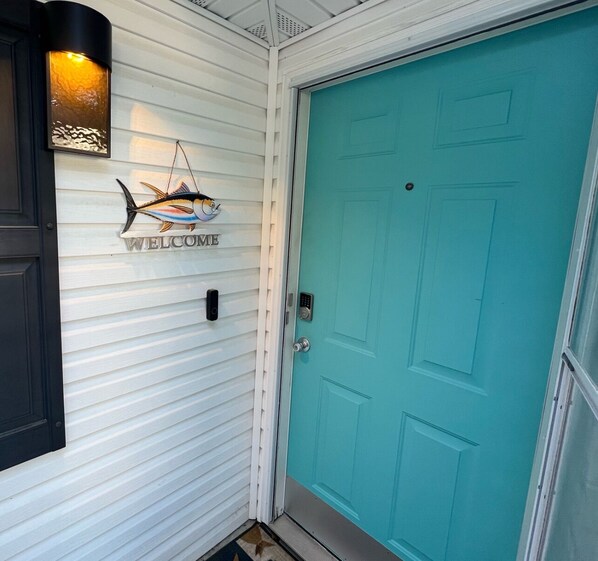 A turquoise door with a 'WELCOME' sign above it, a black mailbox, and a wall-mounted light fixture.