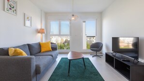 A modern living room with a grey sofa, a green rug, a wooden stool, a TV stand, and a city view through large windows. - Bright, Immaculate apartment in the Estepona port (Estepona)