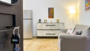A modern living room with a grey sofa, a wooden floor, a white cabinet, and a refrigerator. - Syraka Courtyard - Apartment Ortigia Wifi (Siracusa)