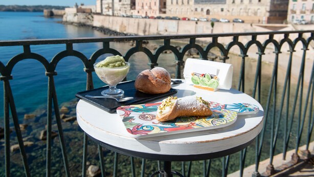 A table with pastries and a dessert on a balcony overlooking a waterfront with historic buildings. - Syraka Courtyard - Apartment Ortigia Wifi (Siracusa)