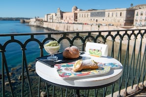 A table with pastries and a dessert on a balcony overlooking a waterfront with historic buildings.