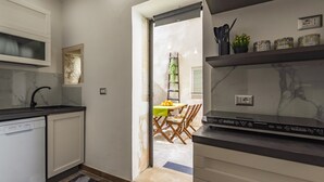 A compact kitchen with white cabinets, a black countertop, and a built-in dishwasher. There is a small window above the sink and a door leading to a dining area with a table and chairs. - Syraka Courtyard - Apartment Ortigia Wifi (Siracusa)