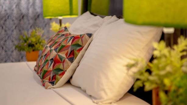 A neatly made bed with white linens and a colorful geometric pillow. There are two green lamps on either side of the bed, and a small potted plant on the nightstand.