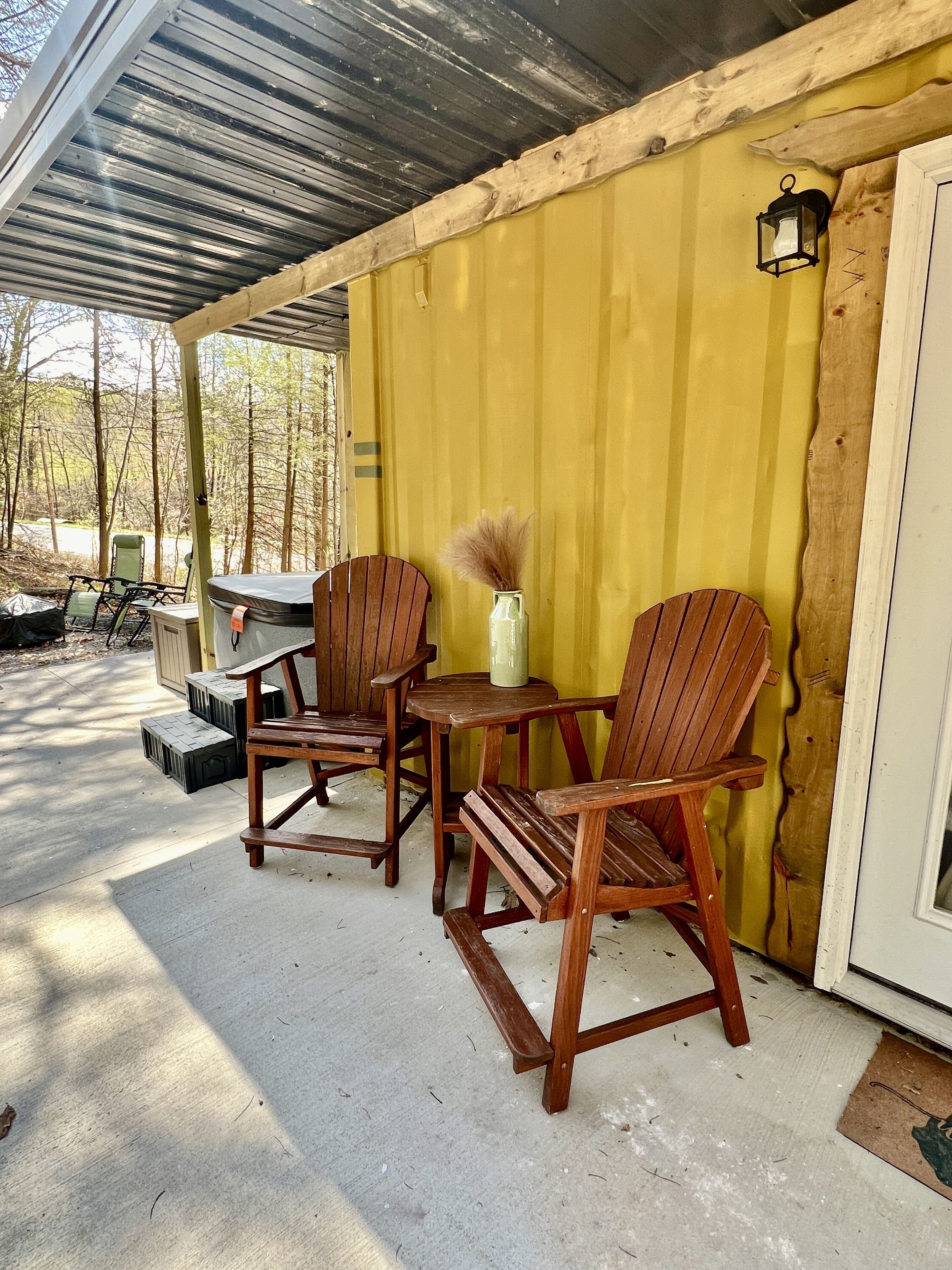 A covered outdoor seating area with wooden chairs and a table, a yellow wall, and a door with a wooden frame.