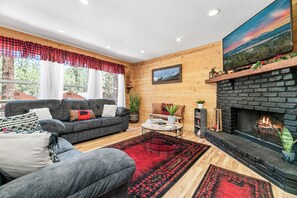 A cozy living room with a fireplace, a sofa set, and a large window with curtains. - The Twisted Twig (Big Bear Lake)