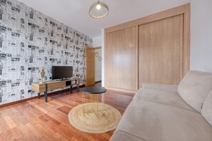 A modern living room with a wooden floor, a sofa, a TV on a wooden console, and a patterned wallpaper.
