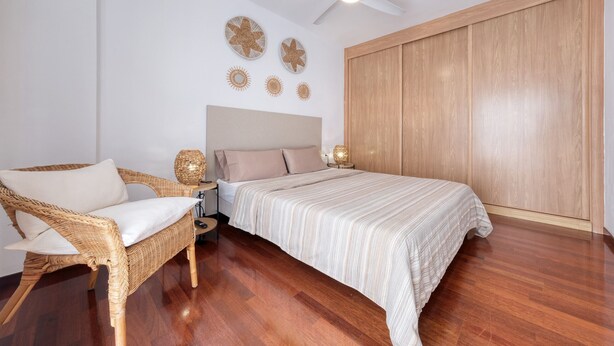 Une chambre à coucher avec un lit, une chaise en osier, un ventilateur de plafond et une armoire en bois.