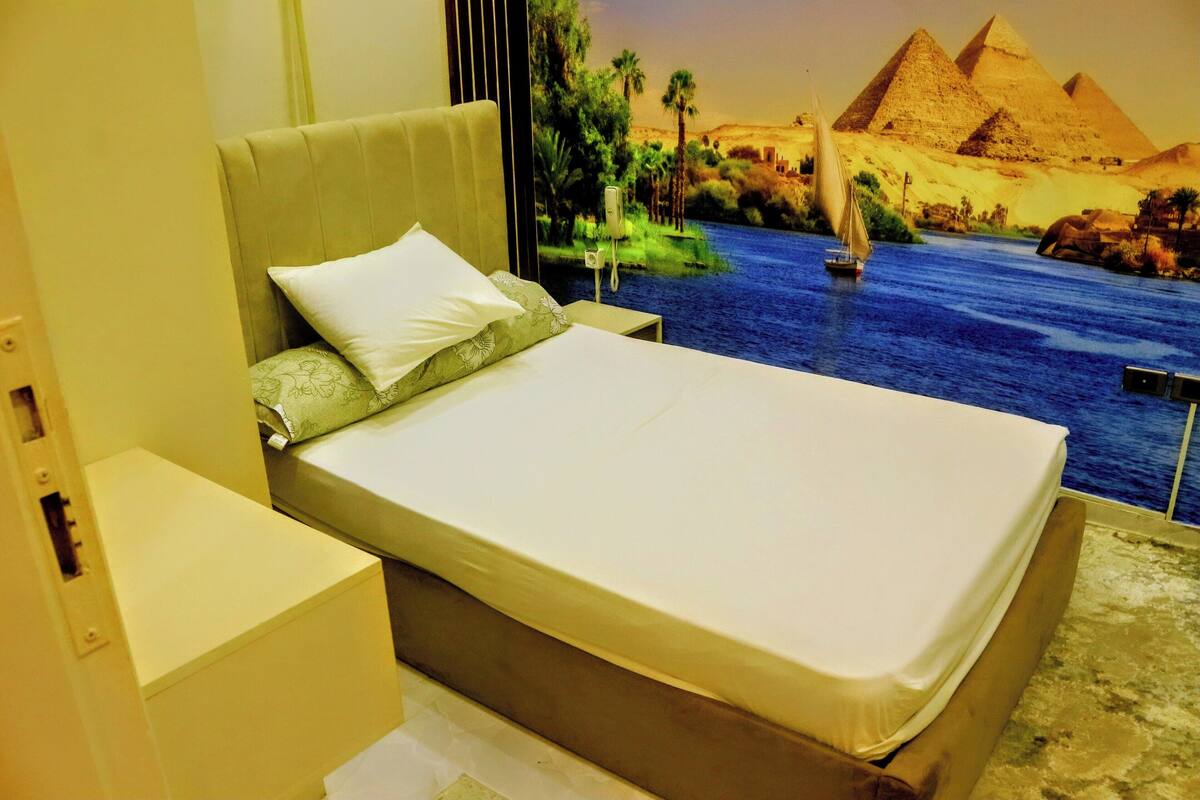 A bed with a view of a mural depicting a desert landscape with pyramids and a river.