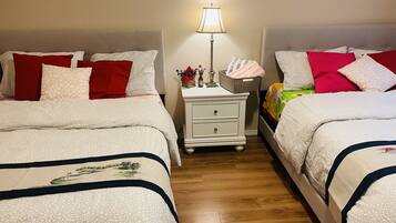 Een hotelkamer met twee bedden, elk voorzien van beddengoed met een patroon en rode kussens. Er is een nachtkastje met een lamp, een mandje en een kleine vaas met bloemen. De kamer heeft houten vloeren en een neutraal kleurenpalet.