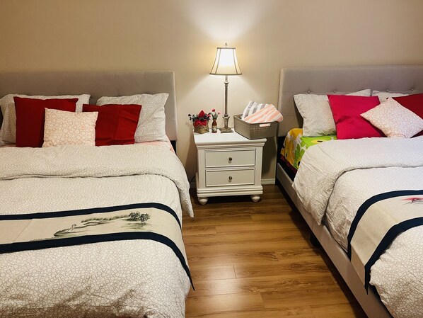 Ein Hotelzimmer mit zwei Betten, jeweils mit gemusterten Betttextilien und roten Kissen. Ein Nachttisch mit Lampe, Korb und kleiner Blumenvase. Das Zimmer hat Holzböden und eine neutrale Farbgestaltung.