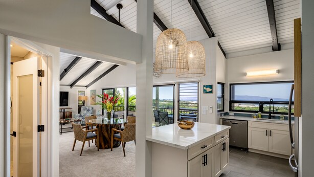 A modern kitchen with a large island, a dining area with a round table and chairs, and a view of the outdoors through large windows. - Upscale and Elegant Vistas (Kihei)