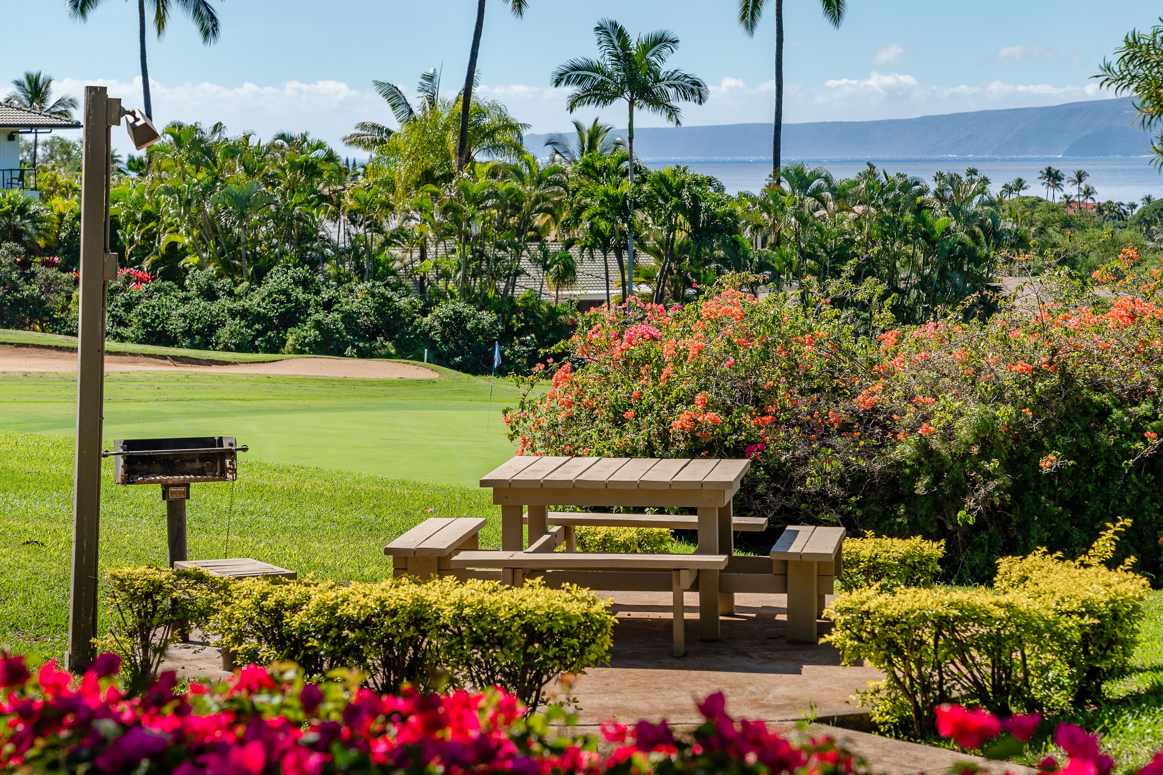 Un campo de golf con una mesa de picnic de madera y bancos, rodeado de vegetación exuberante y plantas en flor.