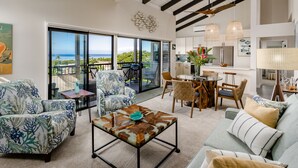A spacious living area with a large sliding glass door leading to a balcony with a view of the ocean. There is a dining area with wicker furniture and a kitchen with modern appliances. - Upscale and Elegant Vistas (Kihei)