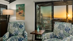 A room with two patterned armchairs, a small wooden table, a painting, and a view of the sunset through a glass door. - Upscale and Elegant Vistas (Kihei)