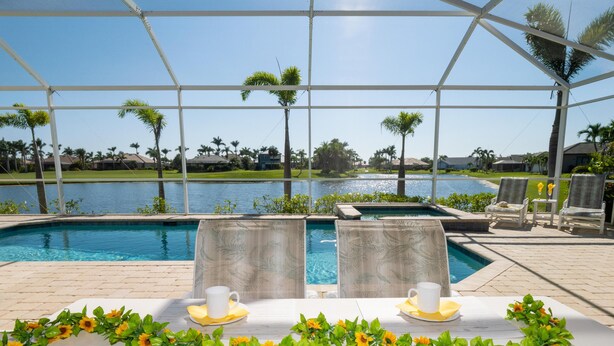 Un espace aménagé au bord de la piscine, avec deux chaises longues, une table décorée de tournesols et une vue sur un lac et des palmiers.