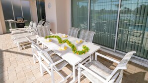 Un espace patio avec une table blanche dressée pour le repas, entourée de chaises blanches, et un barbecue à l’arrière-plan.