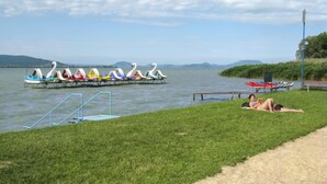 Zona junto al lago con una fila de coloridas lanchas con forma de cisne en un muelle, un área verde con una pequeña piscina y una persona tomando el sol.