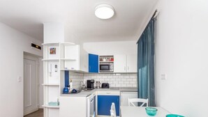 Una cocina compacta con armarios blancos y azules, un microondas, una cafetera y una pequeña zona de comedor con una mesa y sillas blancas.