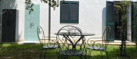 An outdoor seating area with a round table and four chairs, a green umbrella, and a white building with green shutters.