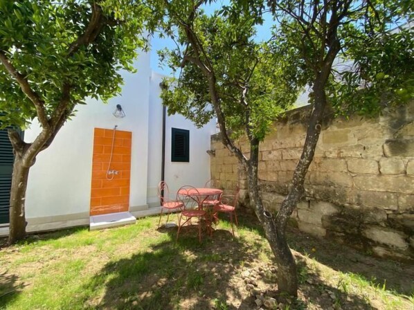 A courtyard with a white building, a brick wall, and a tree. There is a table with chairs and a small window.
