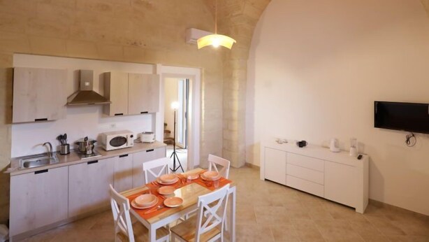 A compact kitchen with white cabinets, a dining table set for four, and a TV mounted on the wall. - Spicchi di Salento - Arancio by Interhome (Vernole)