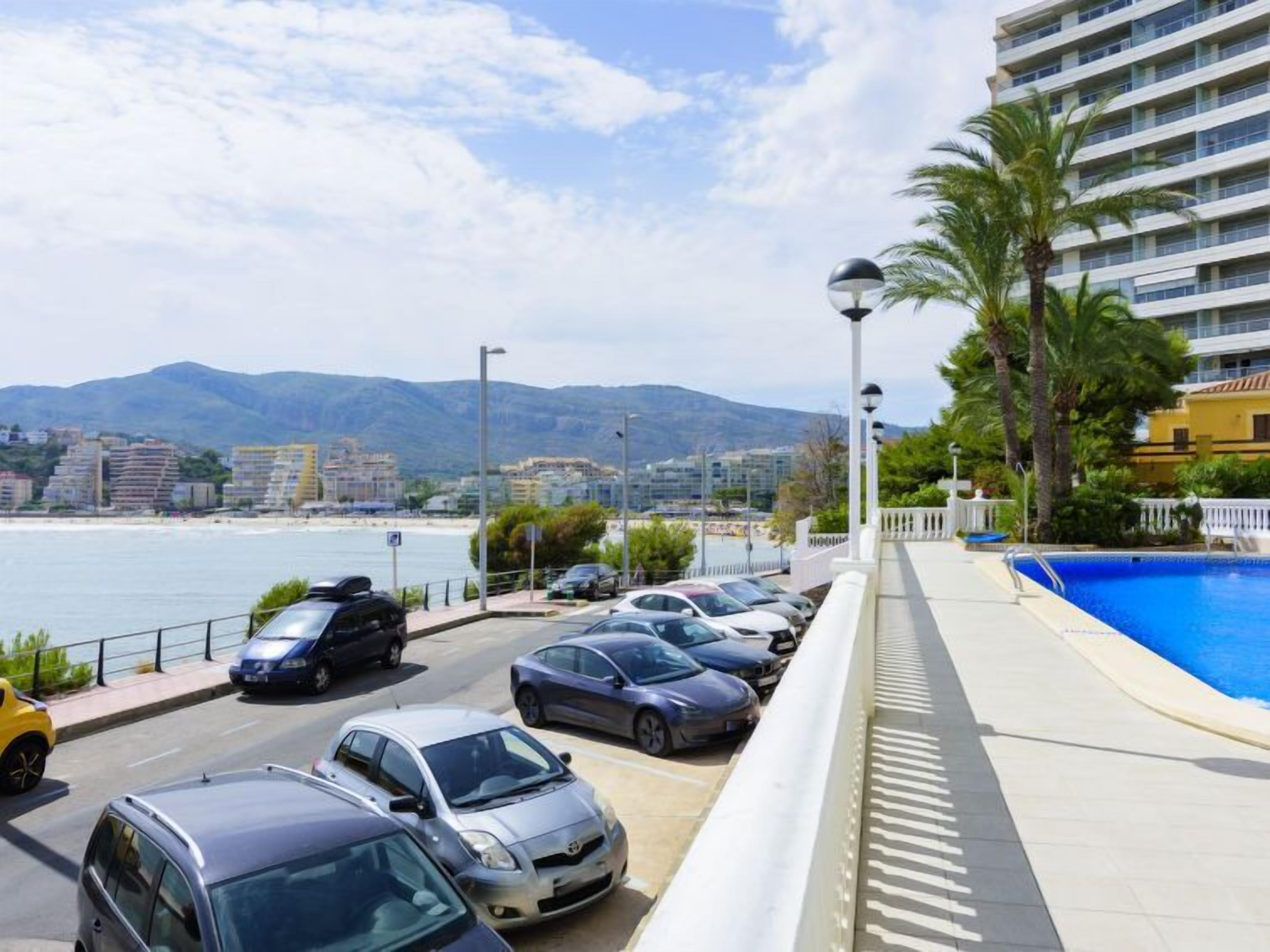 Hotel com piscina, carros estacionados na frente e vista para o mar e montanhas.