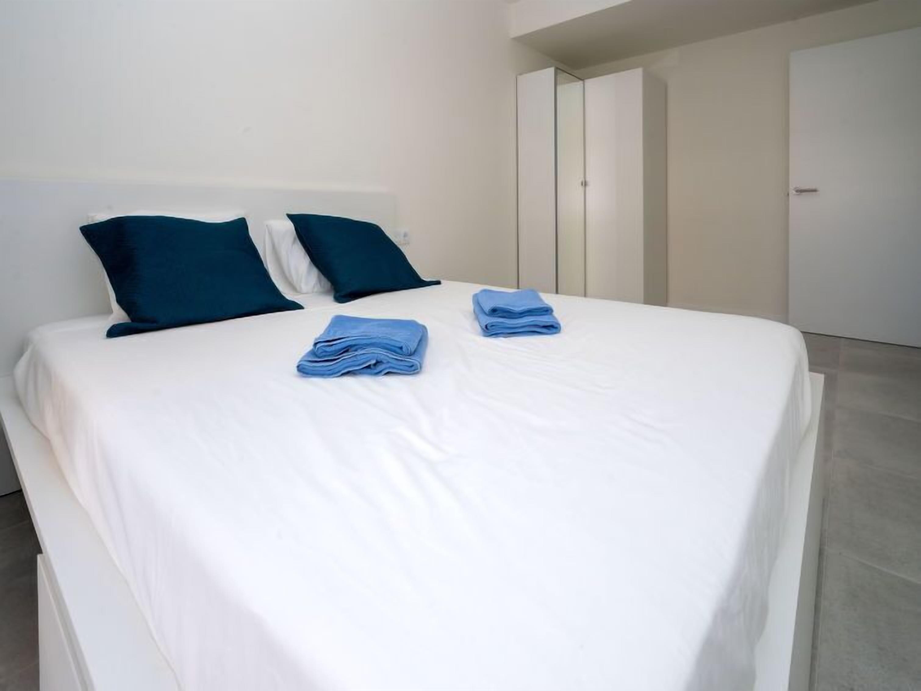 Ein ordentlich bezogenes Bett mit weißen Laken und zwei dunkelblauen Kissen, ein gefaltetes blaues Handtuch auf dem Bett und eine weiße Tür im Hintergrund.