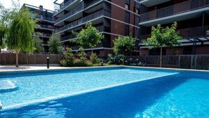 Ein rechteckiger Swimmingpool mit klarem, blauem Wasser, umgeben von einem niedrigen Zaun. Im Hintergrund Hochhäuser mit Balkonen und eine Baumreihe.