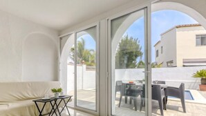 A modern living room with a white sofa, a small table with a plant, and a glass door leading to an outdoor patio with a table and chairs. - Vacation home Luna by Interhome (Empuriabrava)