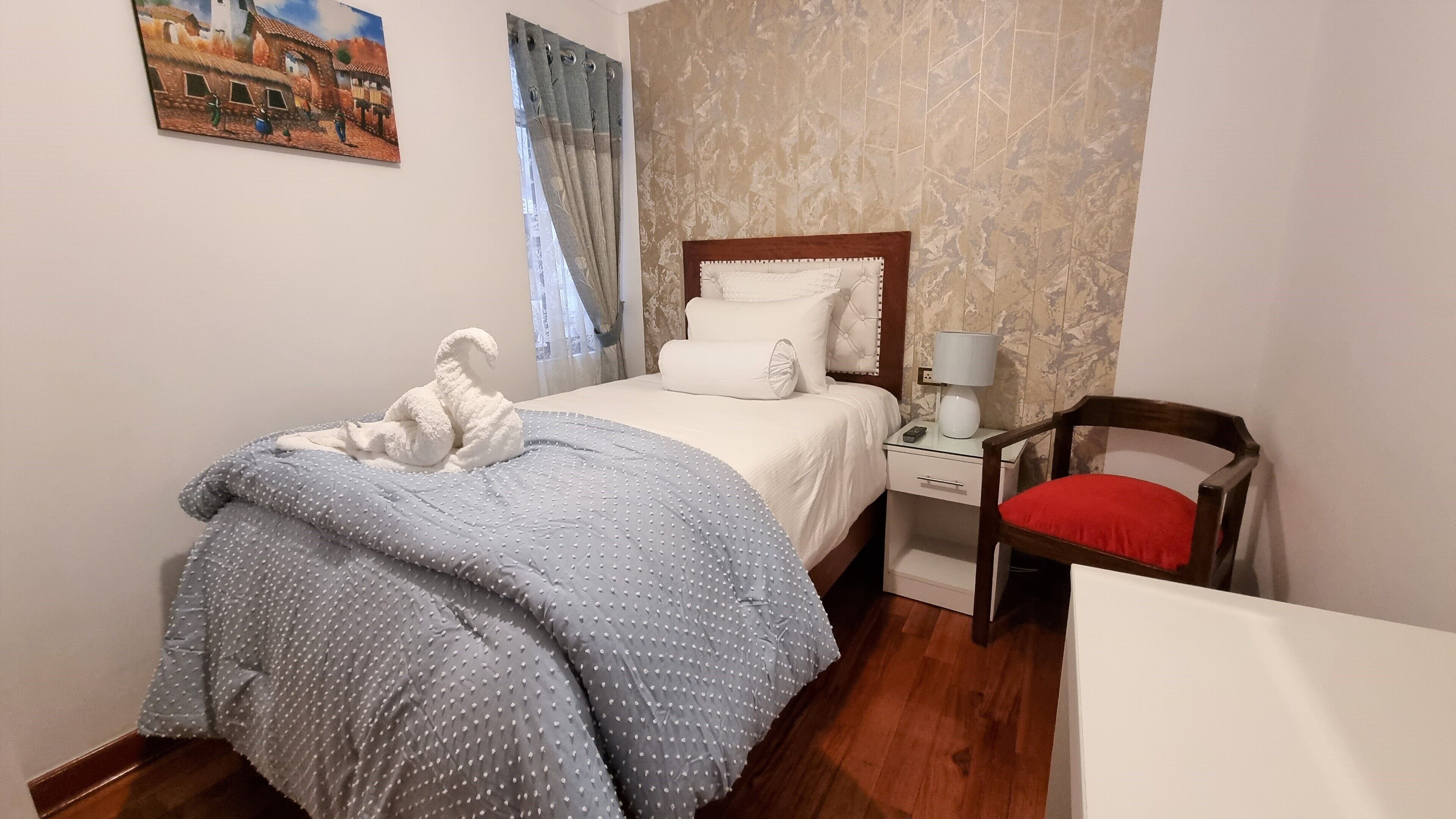 A neatly made bed with a blue polka dot blanket, a wooden nightstand with a lamp, and a red chair.