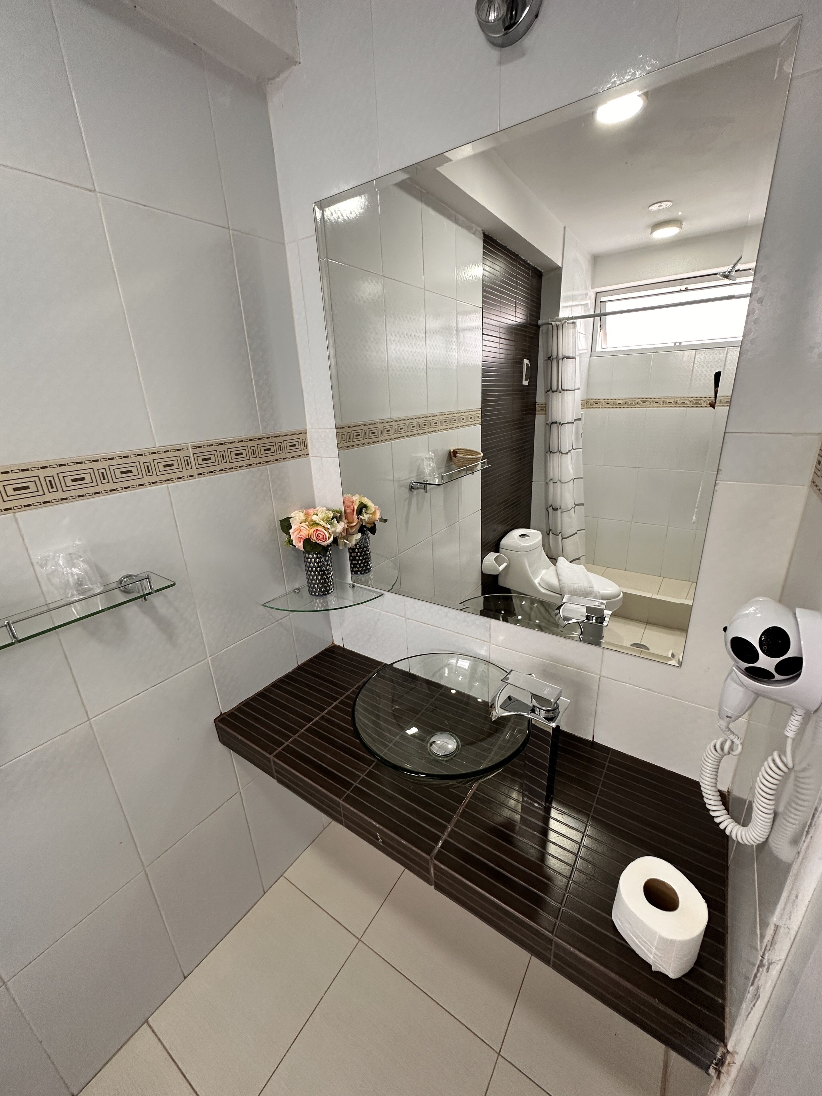Un baño con un lavabo de vidrio, un dispensador de papel higiénico, un secador de pelo y un espejo.