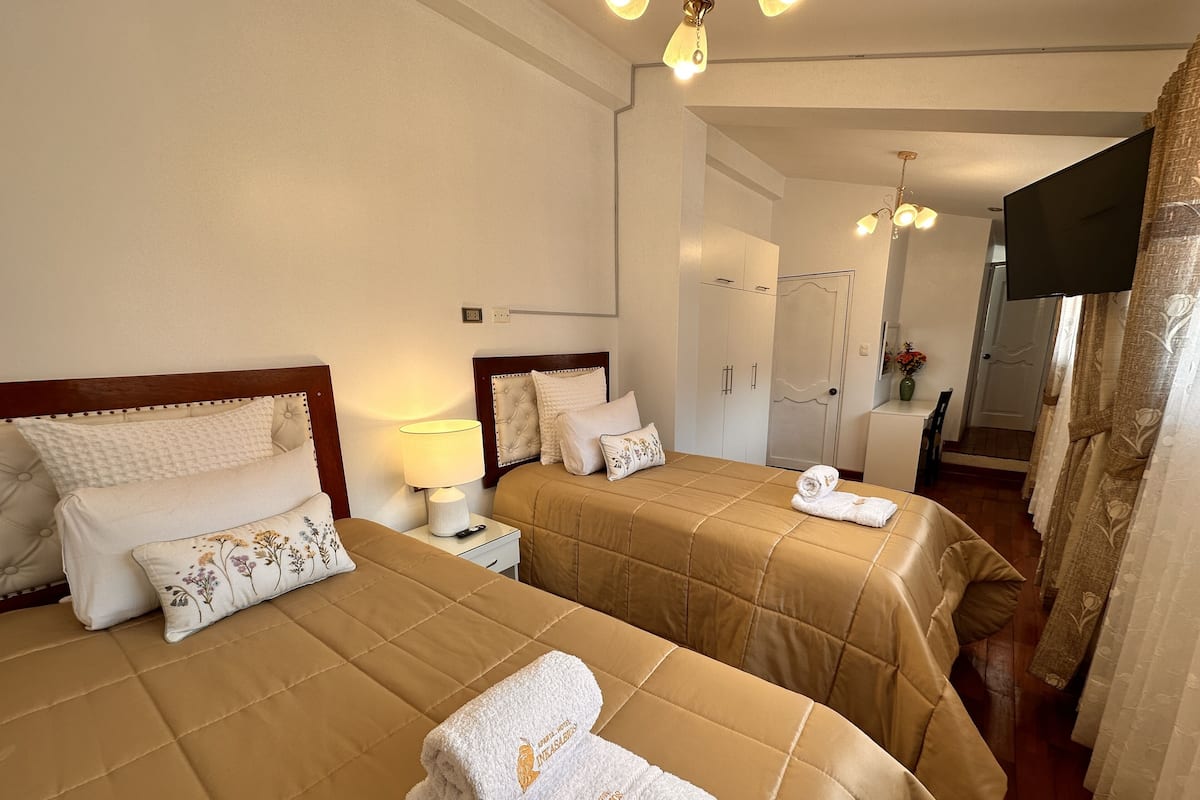 A hotel room with two beds, each with a headboard, bedside tables, and lamps. There is a television on the wall and a door leading to another room.
