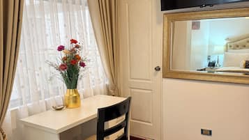 Hotellihuone, jossa on valkoinen työpöytä, tuoli, seinälle asennettu televisio, ikkuna, jossa on verhot, ja kuviollinen matto.