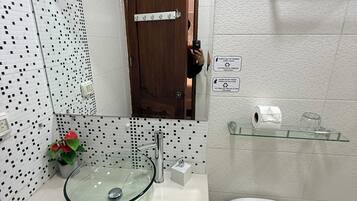 Une salle de bain avec un lavabo en verre, des toilettes, un espace douche et une petite étagère contenant des articles de toilette.
