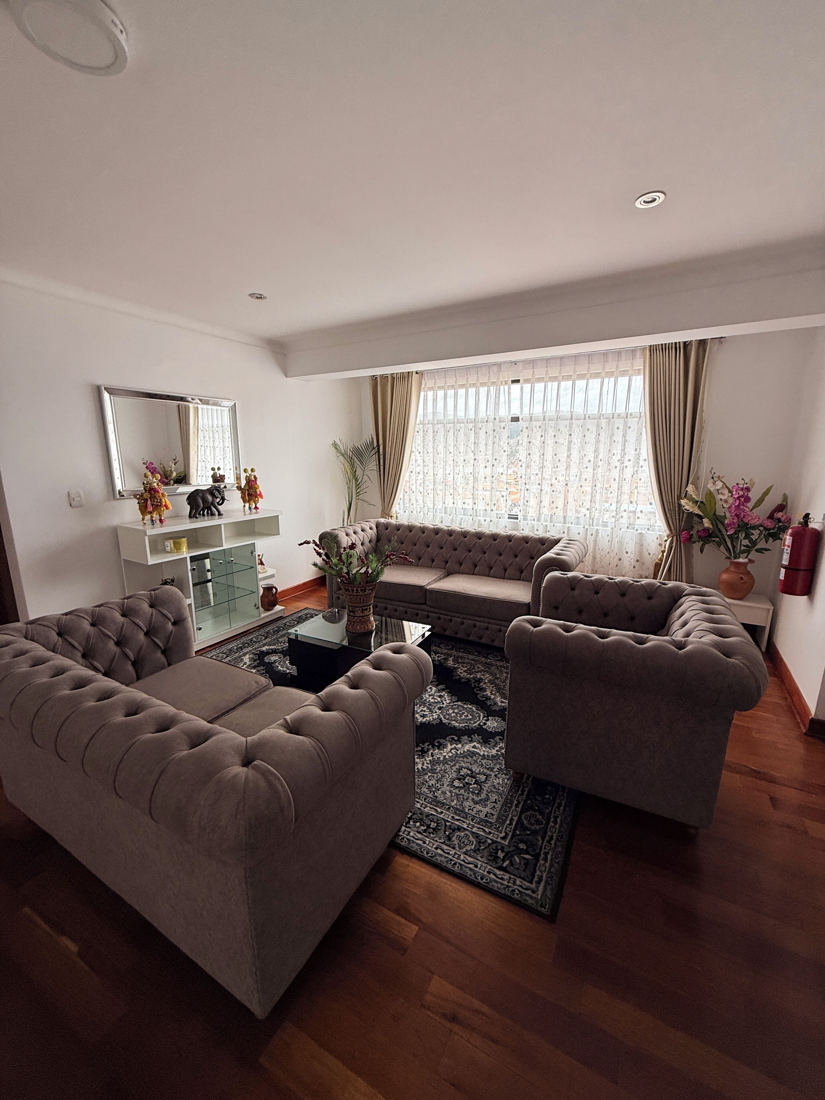 A living room with a sofa, two armchairs, a coffee table, a glass cabinet, and a large window with curtains.