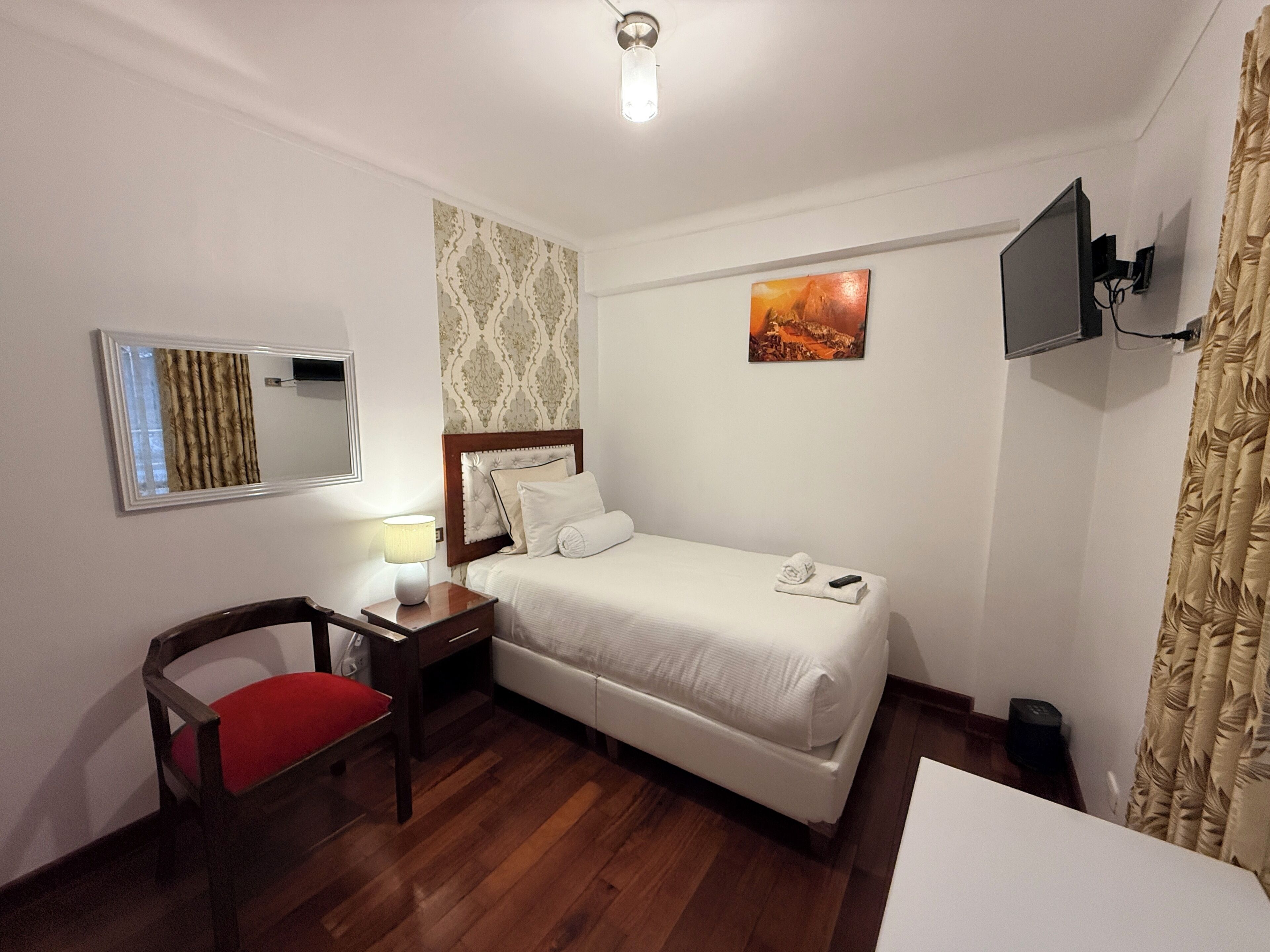 A hotel room with a bed, a desk with a chair, a TV, a painting, and a window with curtains.