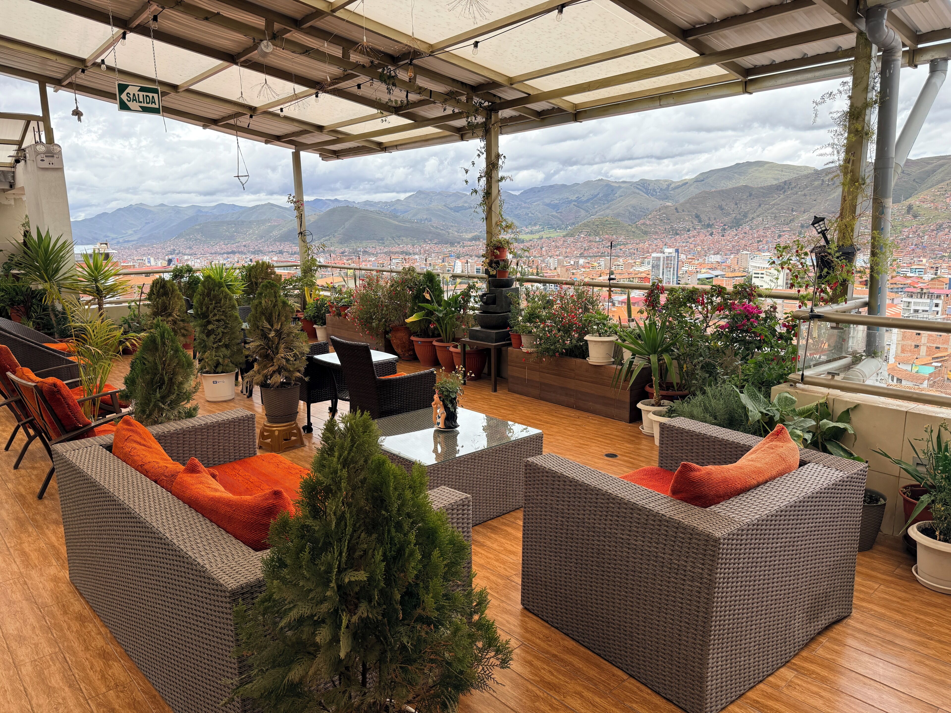 Un espacio de estar al aire libre cubierto, con mobiliario de mimbre, plantas en macetas y vistas a un paisaje urbano y montañas.