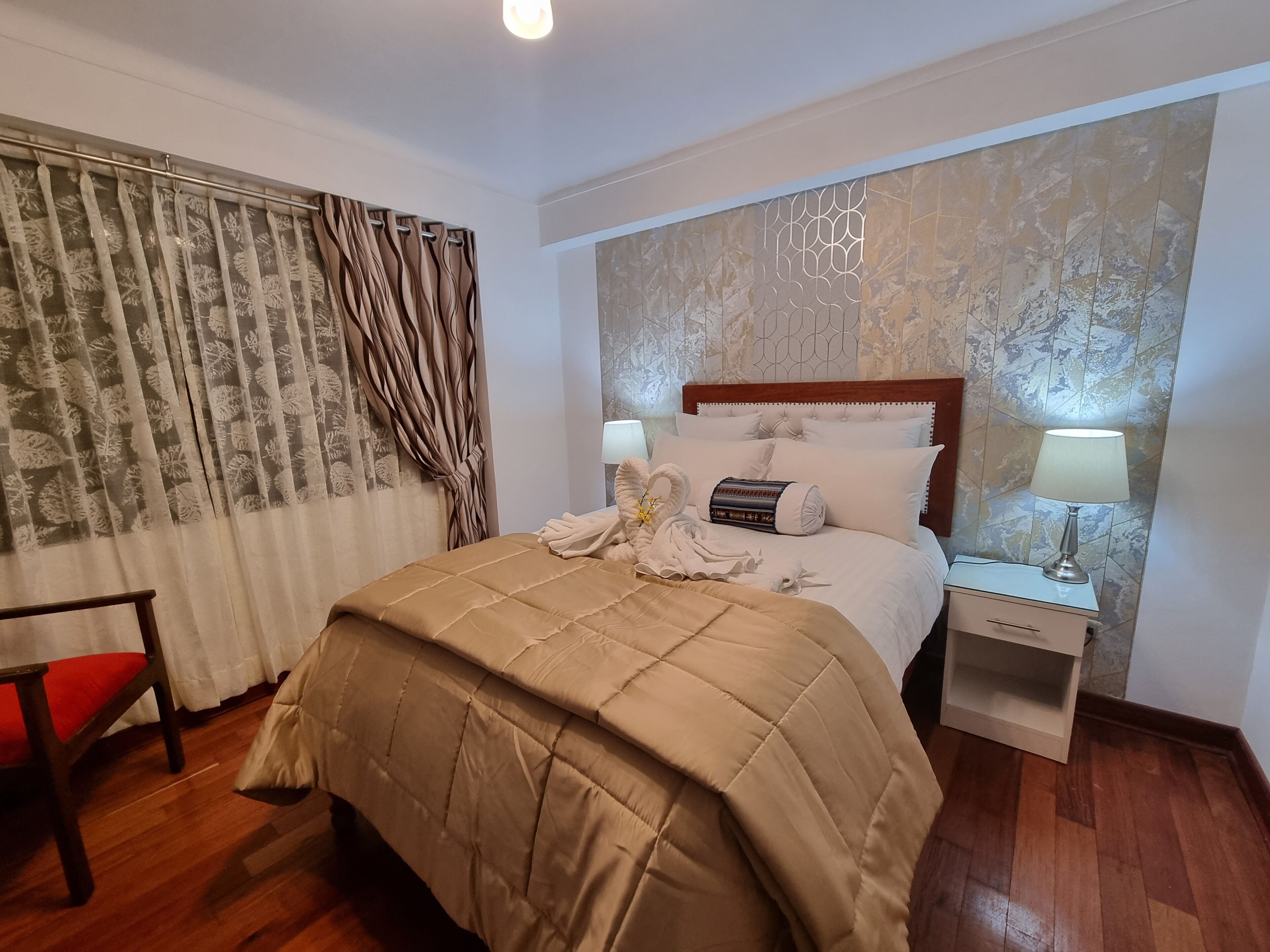 Un lit bien fait, recouvert d’une courtepointe beige, une table de chevet en bois avec une lampe, et une chaise avec un coussin rouge.