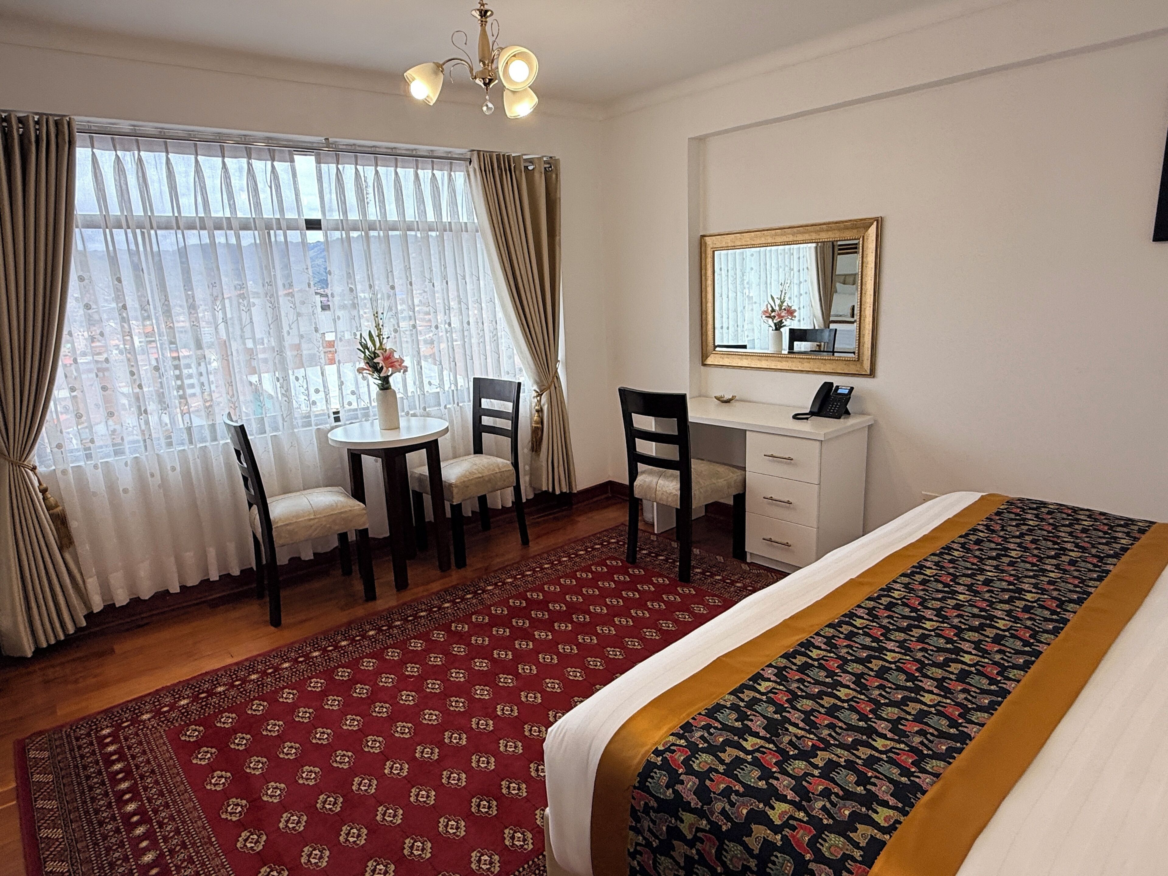 A hotel room with a bed, a desk, a chair, a small table with a vase of flowers, and a large window with curtains.