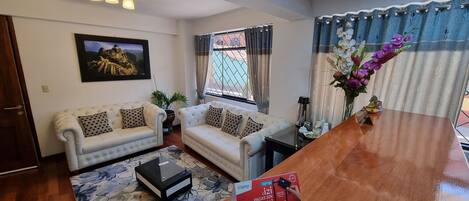 Una sala de estar con dos sofás blancos, una mesa de centro, un televisor y un ventanal con cortinas.