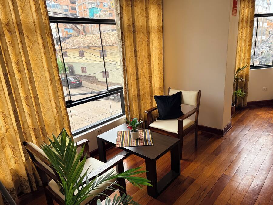 Una habitación con un ventanal grande, piso de madera, una silla con cojín, una mesita con una revista y una planta en maceta.