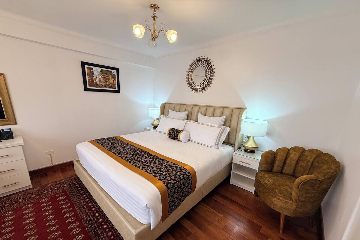 A hotel room with a large bed, bedside tables, a chair, a chandelier, and a wall decoration.