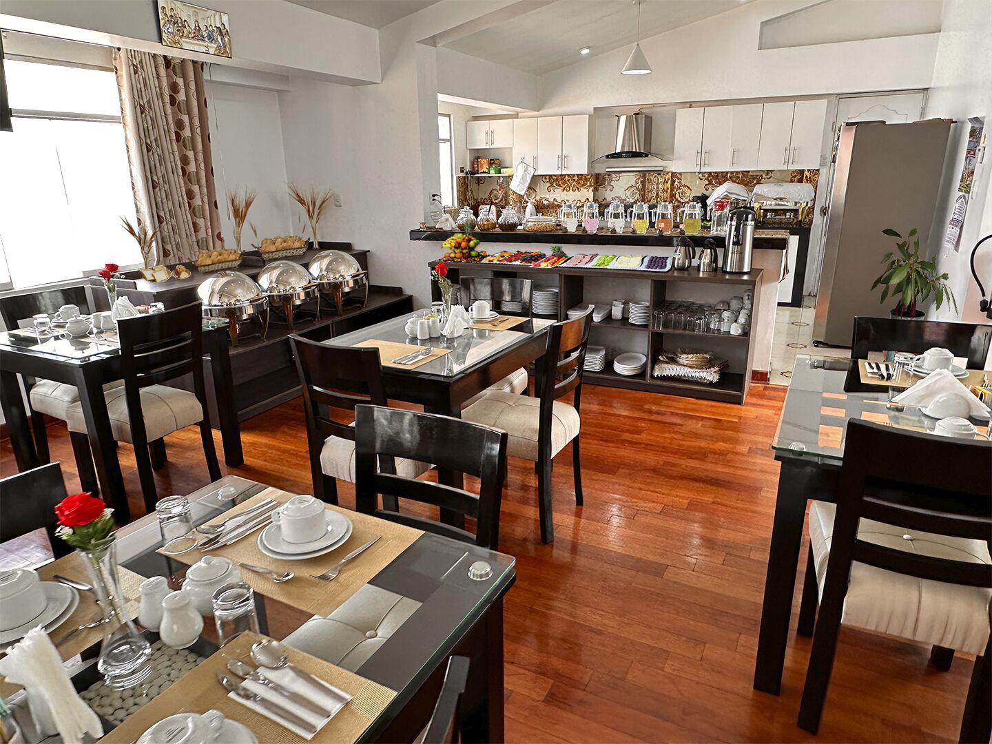 A modern dining area with multiple tables set for service, a kitchen in the background, and a large window with curtains.