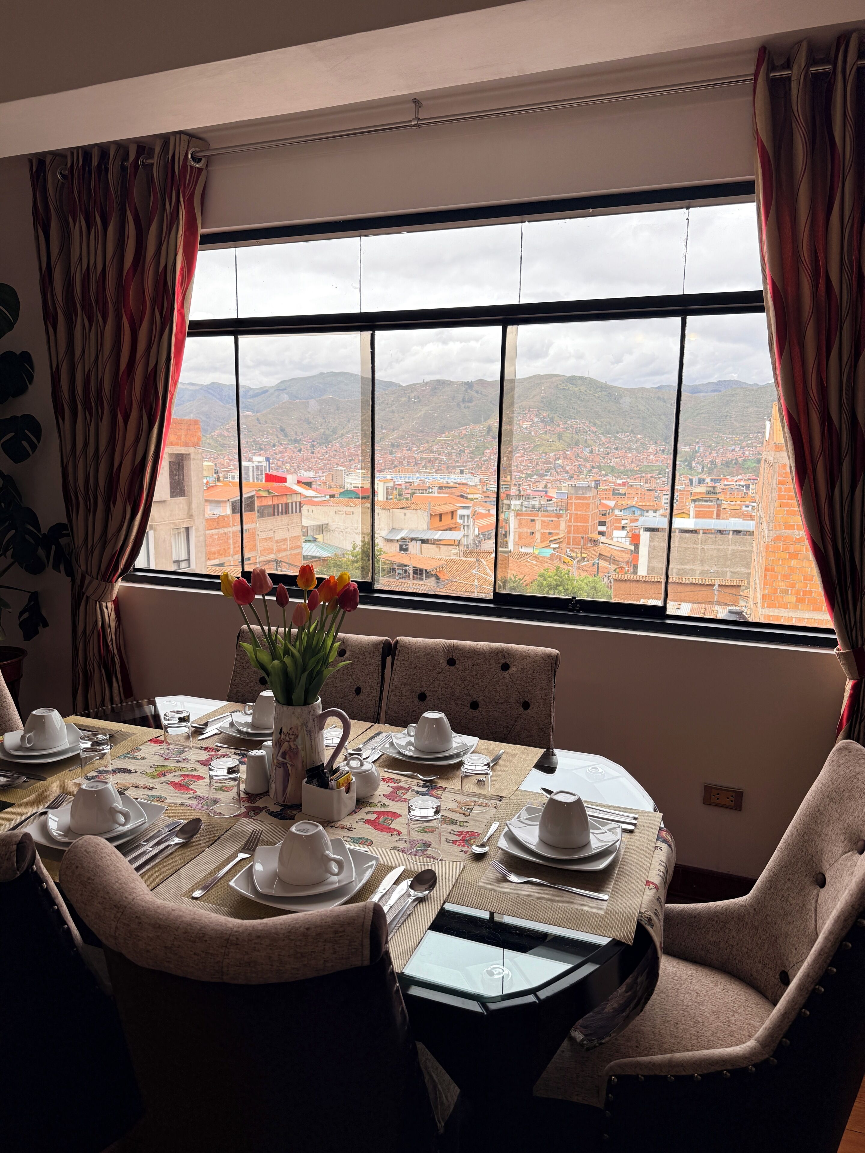 A dining area with a table set for four, a vase with flowers, and a view of a cityscape through a large window.