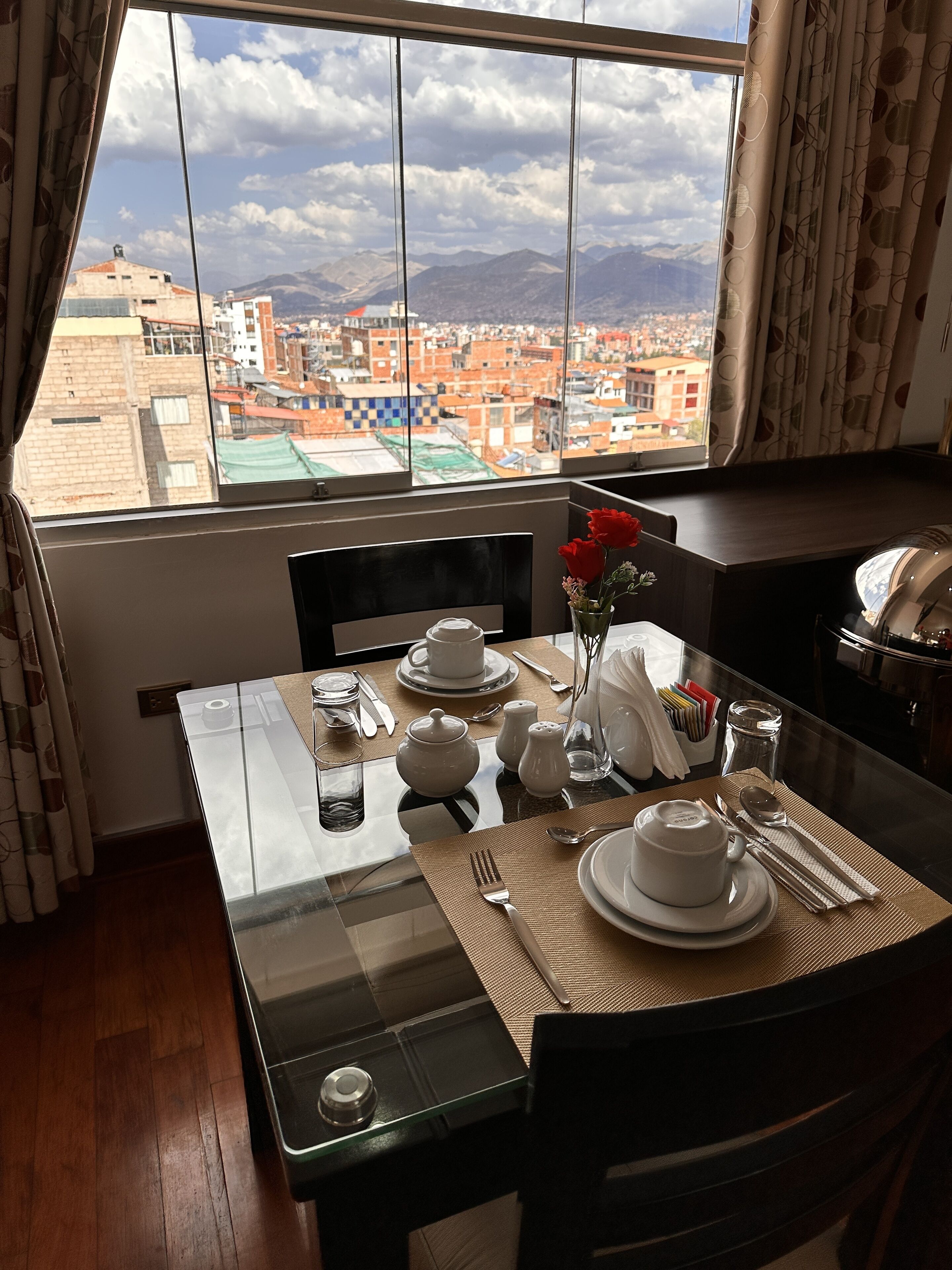 A dining area with a table set for two, a view of a cityscape through a large window, and a vase with red flowers on the table.