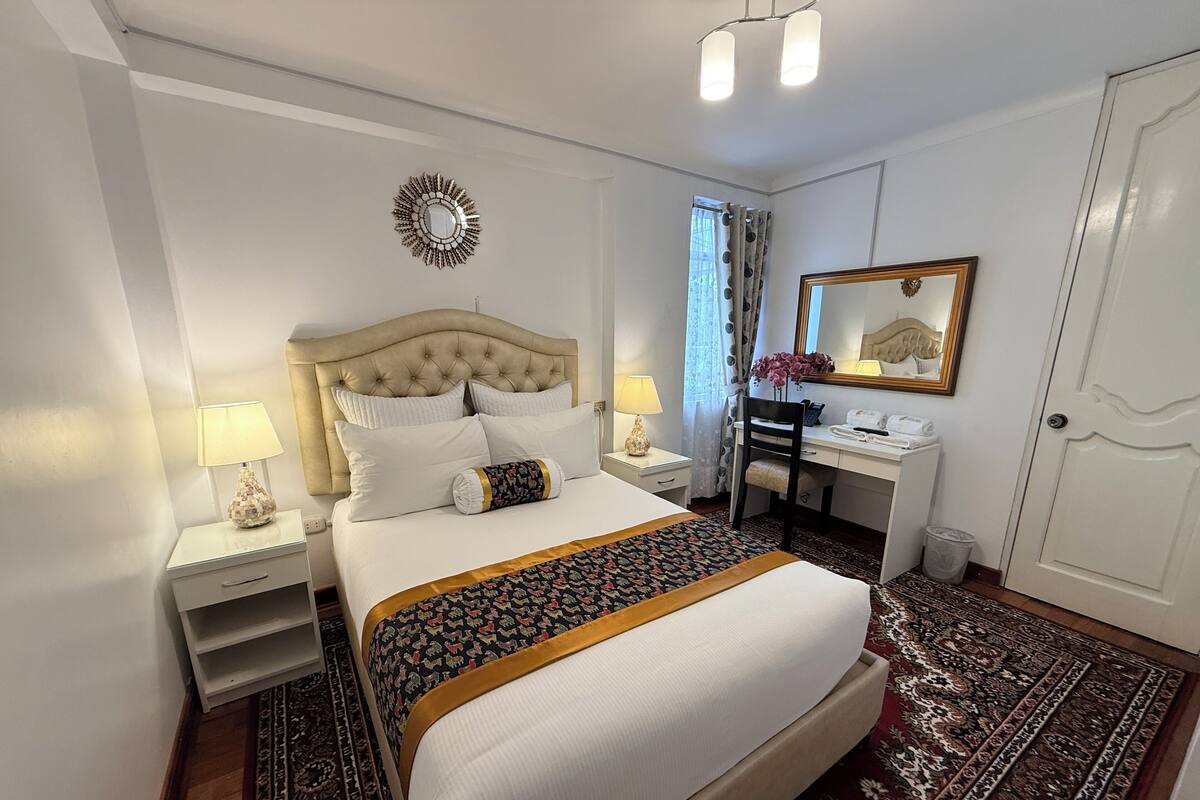 A hotel room with a bed, bedside tables, a desk, a chair, a mirror, and a door.