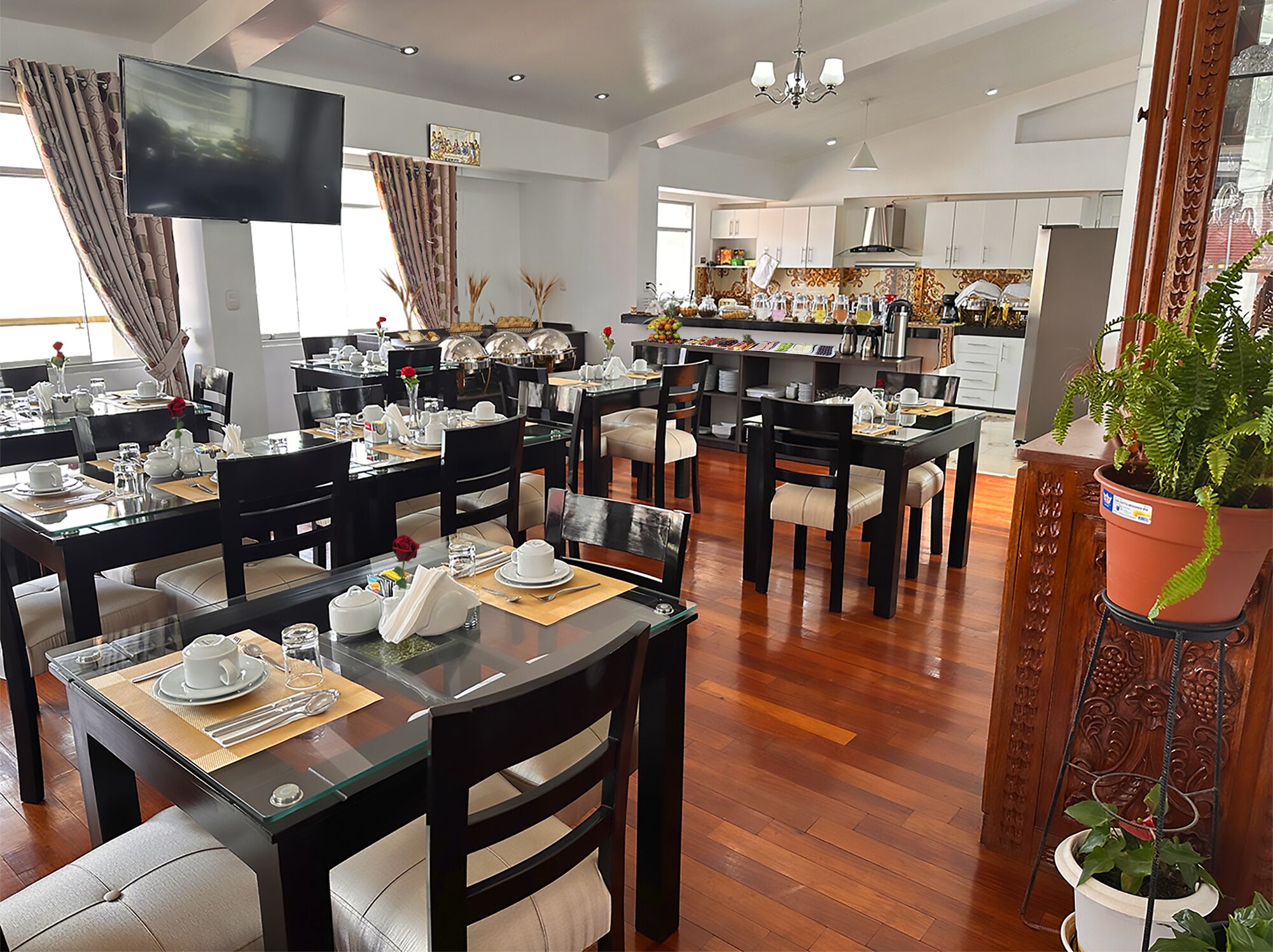 A hotel dining area with multiple tables set for breakfast, a kitchen in the background, and a large flat-screen TV mounted on the wall.