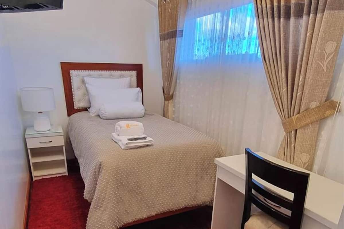 A bedroom with a bed, bedside table, desk, chair, and a TV mounted on the wall.
