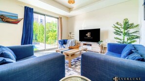 A modern living room with a large TV, blue sofas, a wooden coffee table, and a large window with curtains. - SYCAMORE Damac Hills 2 {Akoya By Damac] (Downtown Dubai)